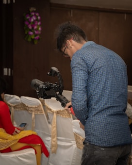 A person in a blue patterned shirt is operating a camera mounted on a stabilizer. They are standing in a room with chairs covered in white cloth, adorned with gold sashes. Another person wearing a red and yellow traditional outfit is seated nearby, looking at a mobile phone.