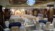 A banquet hall with rows of white chairs adorned with gold sashes and round tables covered with navy blue tablecloths. A large, ornate stage with floral decorations and a luxurious red and gold couch is set up towards the back. The ceiling is decorated with a grand chandelier, and the walls feature intricate wood and floral designs. There are buffet tables on the side with food warmers lined up.