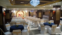 A banquet hall with rows of white chairs adorned with gold sashes and round tables covered with navy blue tablecloths. A large, ornate stage with floral decorations and a luxurious red and gold couch is set up towards the back. The ceiling is decorated with a grand chandelier, and the walls feature intricate wood and floral designs. There are buffet tables on the side with food warmers lined up.