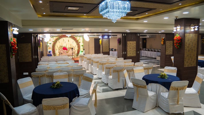 A banquet hall with rows of white chairs adorned with gold sashes and round tables covered with navy blue tablecloths. A large, ornate stage with floral decorations and a luxurious red and gold couch is set up towards the back. The ceiling is decorated with a grand chandelier, and the walls feature intricate wood and floral designs. There are buffet tables on the side with food warmers lined up.