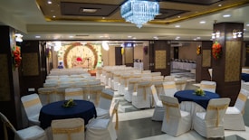 A banquet hall with rows of white chairs adorned with gold sashes and round tables covered with navy blue tablecloths. A large, ornate stage with floral decorations and a luxurious red and gold couch is set up towards the back. The ceiling is decorated with a grand chandelier, and the walls feature intricate wood and floral designs. There are buffet tables on the side with food warmers lined up.