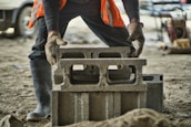 A person wearing gloves and an orange safety vest is in the process of stacking concrete cinder blocks on a construction site. The image also shows the person wearing black pants and rubber boots. The background contains construction equipment and a vehicle.