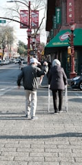 An elderly couple is walking down a city street. One person uses a walker, while the other raises a hand in a gesture. The street is lined with red lanterns and banners, and there are shops and cars visible in the background.