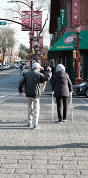 An elderly couple is walking down a city street. One person uses a walker, while the other raises a hand in a gesture. The street is lined with red lanterns and banners, and there are shops and cars visible in the background.