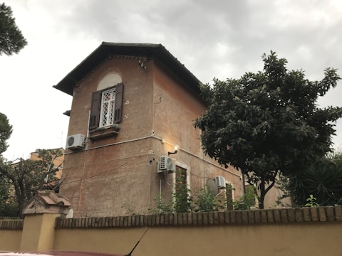 A two-story house with a rustic appearance, featuring brown walls and several air conditioning units attached to its exterior. There is a window with shutters on the second floor and small trees and plants surrounding the building. The sky appears overcast.