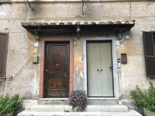 Two adjacent doorways with different styles on a textured, weathered wall. The left door is dark brown with a brick frame, while the right door is light gray and plainer. Above the doors, a small awning with curved tiles provides some cover. Beside each door are elements such as a mailbox, doorbell, and house numbers, 32 and 30. Greenery with small flowers is at the base of the wall, adding a touch of nature.