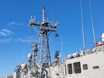 A naval ship is depicted with a complex array of antennas and radar equipment on its mast. The sky is clear with only a few clouds, providing a backdrop to the intricate structure. A flag is visible among the equipment, and there are several spherical and cylindrical elements mounted on the ship