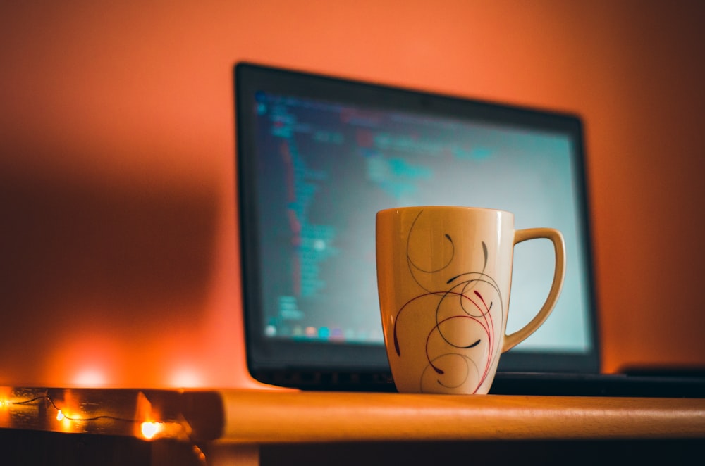 Una taza de café sentada en un escritorio junto a una computadora portátil