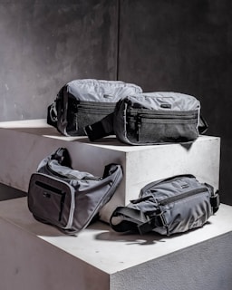 Four grey bags are arranged on a pair of concrete blocks. The bags have minimalistic design features, including black zippers and straps.