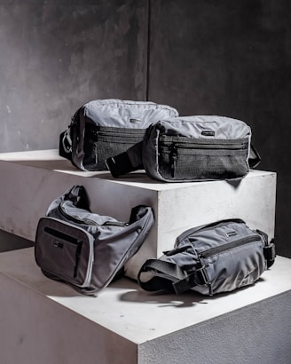 Four grey bags are arranged on a pair of concrete blocks. The bags have minimalistic design features, including black zippers and straps.