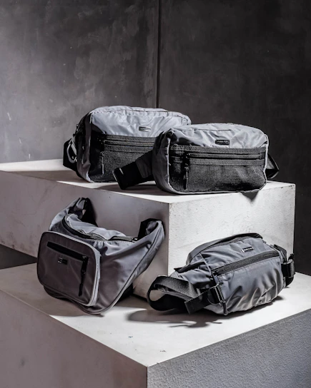 Four grey bags are arranged on a pair of concrete blocks. The bags have minimalistic design features, including black zippers and straps.