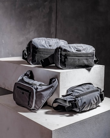 Four grey bags are arranged on a pair of concrete blocks. The bags have minimalistic design features, including black zippers and straps.