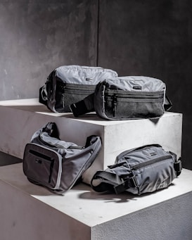 Four grey bags are arranged on a pair of concrete blocks. The bags have minimalistic design features, including black zippers and straps.
