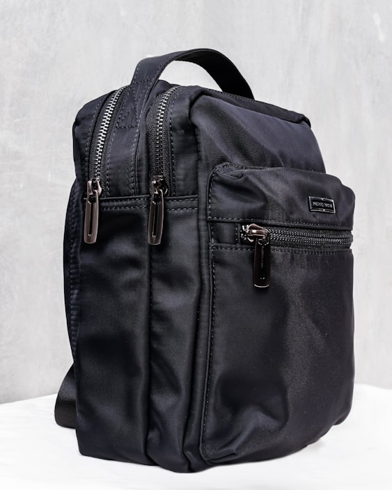 A sleek black backpack with multiple zippered compartments and a sturdy carrying handle. The design is minimalist and practical, suggesting functionality and durability.