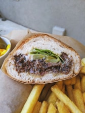 A sandwich filled with shredded meat and fresh greens is placed on a piece of brown paper. Beside the sandwich, there is a serving of crispy golden French fries. In the background, there is a small bowl containing pickles.