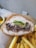 A sandwich filled with shredded meat and fresh greens is placed on a piece of brown paper. Beside the sandwich, there is a serving of crispy golden French fries. In the background, there is a small bowl containing pickles.