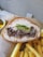 A sandwich filled with shredded meat and fresh greens is placed on a piece of brown paper. Beside the sandwich, there is a serving of crispy golden French fries. In the background, there is a small bowl containing pickles.
