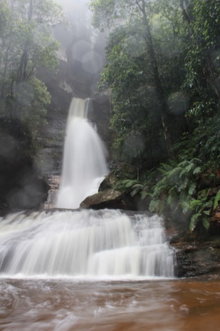A multi-tiered waterfall flowing into a pond with soft lighting highlighting the textured rocks at dusk.