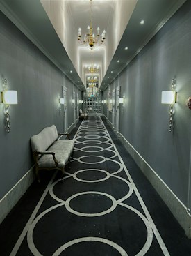 A long, elegantly decorated hallway with gray walls and a dark carpet featuring circular patterns. The ceiling is adorned with ornate chandeliers that provide warm lighting. Wall sconces line the sides, adding to the sophisticated ambiance. A vintage-style sofa sits on the left side of the corridor.