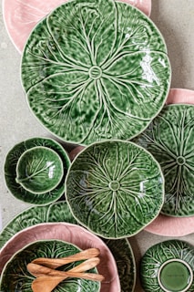 A collection of ceramic plates and bowls with a leafy design in green and pink hues is arranged on a flat surface. The ceramics are complemented by a couple of small wooden spoons. The designs mimic the texture and appearance of leaves, creating a natural and organic aesthetic.