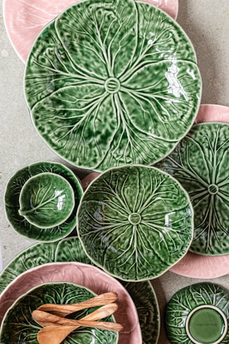 A collection of ceramic plates and bowls with a leafy design in green and pink hues is arranged on a flat surface. The ceramics are complemented by a couple of small wooden spoons. The designs mimic the texture and appearance of leaves, creating a natural and organic aesthetic.