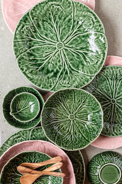 A collection of ceramic plates and bowls with a leafy design in green and pink hues is arranged on a flat surface. The ceramics are complemented by a couple of small wooden spoons. The designs mimic the texture and appearance of leaves, creating a natural and organic aesthetic.