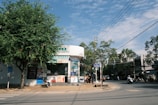 A street corner with a pharmacy or small store with a glass façade and various advertisements on the exterior. There are motorbikes parked in front of the store, and people are riding motorbikes on the street. A large tree provides shade on the left, and there is a clear blue sky with scattered clouds overhead.