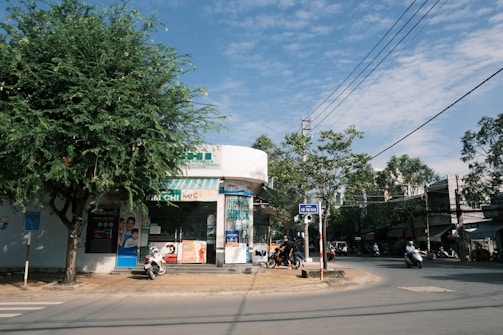 A street corner with a pharmacy or small store with a glass façade and various advertisements on the exterior. There are motorbikes parked in front of the store, and people are riding motorbikes on the street. A large tree provides shade on the left, and there is a clear blue sky with scattered clouds overhead.