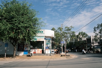 A street corner with a pharmacy or small store with a glass façade and various advertisements on the exterior. There are motorbikes parked in front of the store, and people are riding motorbikes on the street. A large tree provides shade on the left, and there is a clear blue sky with scattered clouds overhead.