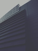 A modern building with a geometric design, featuring dark horizontal slats and a grid-like window structure. The architectural style conveys a sense of minimalism and sleekness.