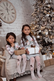 Two children sitting on a cozy sofa holding wrapped gifts. A decorated Christmas tree with lights and ornaments stands next to them. A large, vintage clock hangs on a brick wall behind.