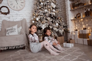 Two children are sitting on the floor in a cozy, decorated room. A snowy Christmas tree with ornaments is in the background. There are several wrapped gifts on the floor beside them. A cozy chair with a 'good vibes' pillow, adorned with a blanket, sits against a white brick wall with a clock and a wreath. The scene is warmly lit with soft, yellow lights.