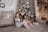 Two children are sitting on the floor in a cozy, decorated room. A snowy Christmas tree with ornaments is in the background. There are several wrapped gifts on the floor beside them. A cozy chair with a 'good vibes' pillow, adorned with a blanket, sits against a white brick wall with a clock and a wreath. The scene is warmly lit with soft, yellow lights.