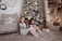 Two children are sitting on the floor in a cozy, decorated room. A snowy Christmas tree with ornaments is in the background. There are several wrapped gifts on the floor beside them. A cozy chair with a 'good vibes' pillow, adorned with a blanket, sits against a white brick wall with a clock and a wreath. The scene is warmly lit with soft, yellow lights.