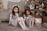Two young children are sitting on a cozy, beige carpet in front of a beautifully decorated Christmas tree with lights. They are wearing matching festive pajamas with cartoon characters on the tops and leggings. Behind them is a comfortable chair draped with a cozy blanket and a decorative pillow that has the words 'Good Vibes' printed on it. Present boxes wrapped in brown paper are arranged around the tree, adding to the festive atmosphere.