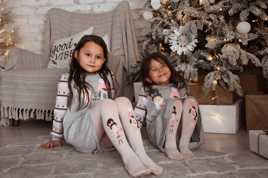 Two young children are sitting on a cozy, beige carpet in front of a beautifully decorated Christmas tree with lights. They are wearing matching festive pajamas with cartoon characters on the tops and leggings. Behind them is a comfortable chair draped with a cozy blanket and a decorative pillow that has the words 'Good Vibes' printed on it. Present boxes wrapped in brown paper are arranged around the tree, adding to the festive atmosphere.