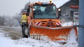 A large orange snowplow is clearing snow from a road surrounded by a snowy landscape. The vehicle has a prominent bright orange plow at the front, and there is light snow on the ground and trees. Road signs and a building with signage are visible in the background.