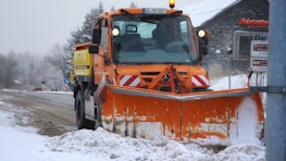A large orange snowplow is clearing snow from a road surrounded by a snowy landscape. The vehicle has a prominent bright orange plow at the front, and there is light snow on the ground and trees. Road signs and a building with signage are visible in the background.