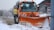 A large orange snowplow is clearing snow from a road surrounded by a snowy landscape. The vehicle has a prominent bright orange plow at the front, and there is light snow on the ground and trees. Road signs and a building with signage are visible in the background.
