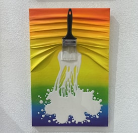 A creative artwork featuring a brush with white paint splattering out from the bristles, set against a textured rainbow background. The colors transition smoothly from orange at the top through yellow, green, and blue shades.