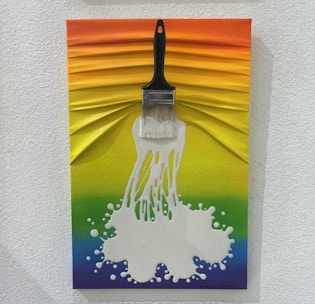 A creative artwork featuring a brush with white paint splattering out from the bristles, set against a textured rainbow background. The colors transition smoothly from orange at the top through yellow, green, and blue shades.