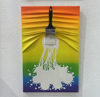 A creative artwork featuring a brush with white paint splattering out from the bristles, set against a textured rainbow background. The colors transition smoothly from orange at the top through yellow, green, and blue shades.