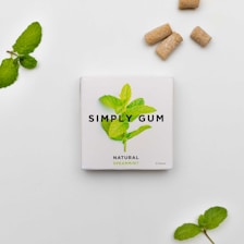 A package of Simply Gum with a spearmint leaf illustration centered on a white background. The package is accompanied by mint leaves and several brownish gum pieces nearby, creating a natural and fresh aesthetic.