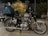 A classic motorcycle parked outdoors with a large blue duffel bag and a helmet on the back seat. There is a plant decorated with small lights in the background, and buildings are partially visible.