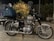 A classic motorcycle parked outdoors with a large blue duffel bag and a helmet on the back seat. There is a plant decorated with small lights in the background, and buildings are partially visible.
