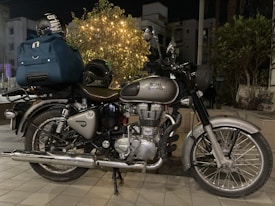 A classic motorcycle parked outdoors with a large blue duffel bag and a helmet on the back seat. There is a plant decorated with small lights in the background, and buildings are partially visible.
