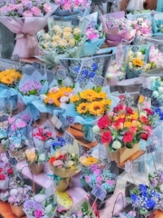 Various bouquets of colorful flowers are neatly arranged on display, each wrapped in decorative paper and labeled with price tags. The assortment includes roses, sunflowers, and other vibrant blooms, set against a wooden surface.