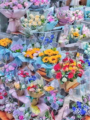 Various bouquets of colorful flowers are neatly arranged on display, each wrapped in decorative paper and labeled with price tags. The assortment includes roses, sunflowers, and other vibrant blooms, set against a wooden surface.