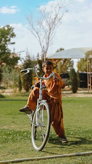 A young boy wearing traditional clothing is joyfully riding a bicycle on a grassy field. In the background, there are some trees and a partially visible structure, possibly a gazebo, under a clear blue sky.