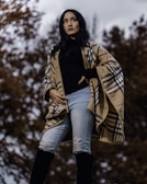 A person standing outdoors wearing a stylish plaid poncho, black top, light blue ripped jeans, and knee-high black boots. The background features trees with autumn leaves and an overcast sky, creating a moody atmosphere.